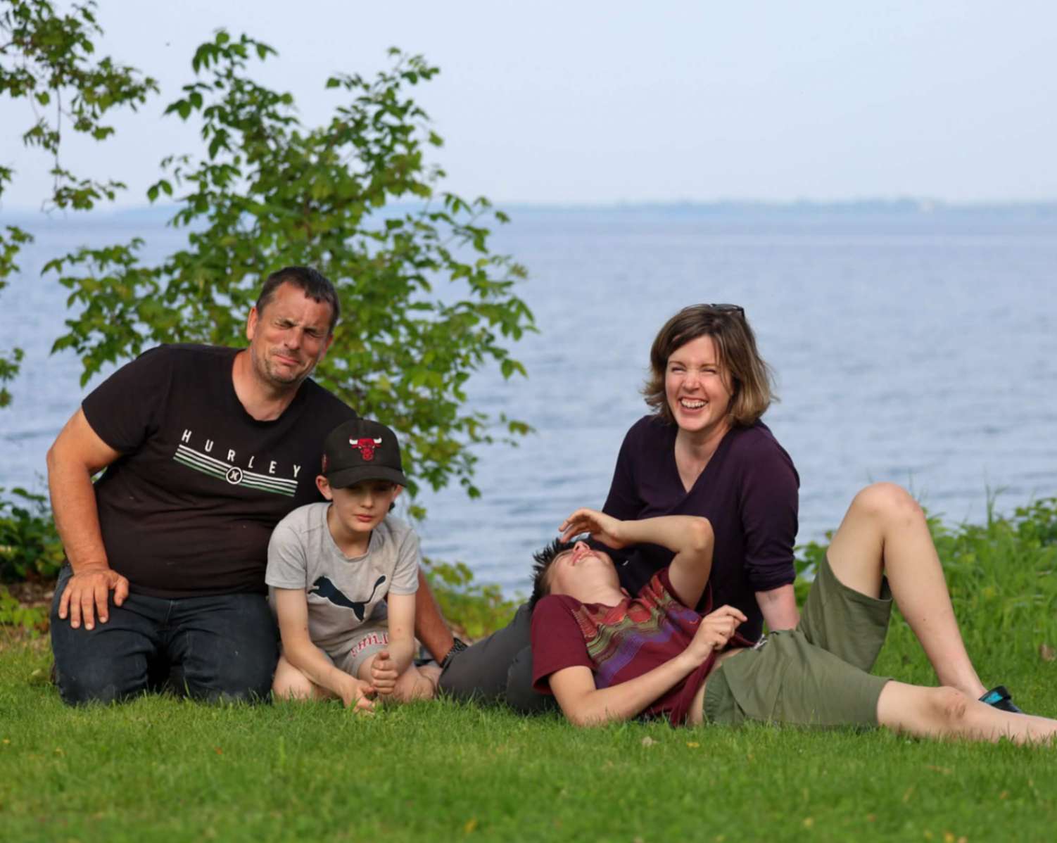 Kent family sitting together on the grass by the water in Eastern Ontario